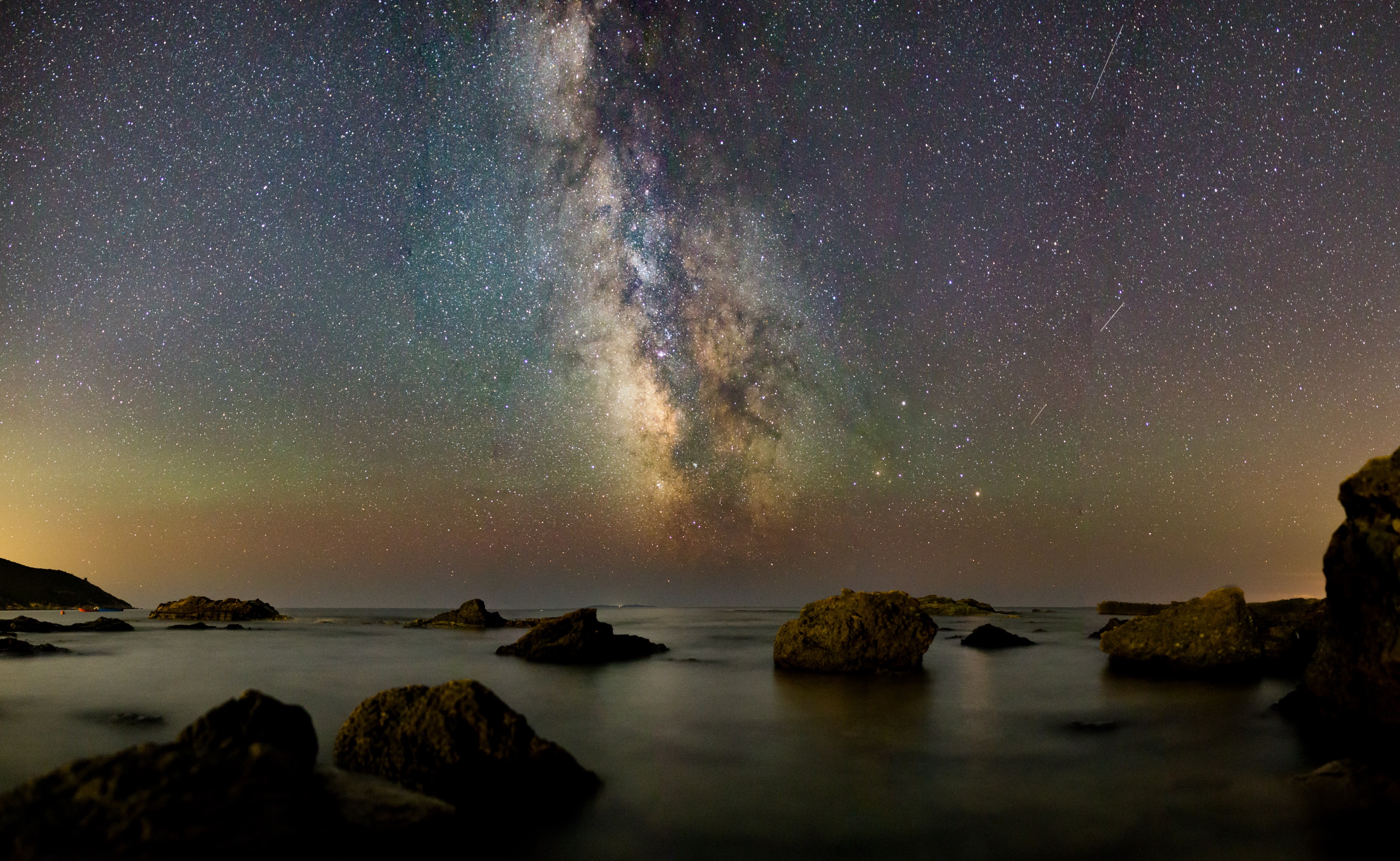 photo of night sky with stars with some rainbow colors above the sea with boulders